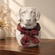 Chalk white dog portrait urn wearing a tartan collar and bow on a dark walnut sideboard.