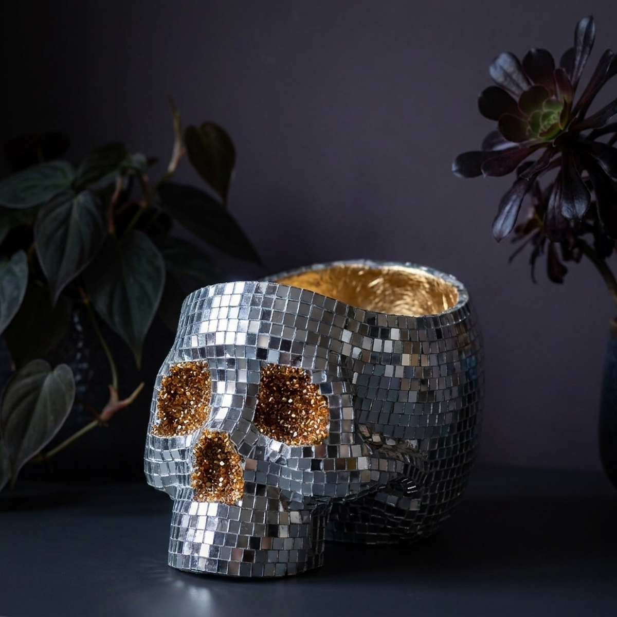 Silver disco skull planter with gold eye sockets beside dark foliage