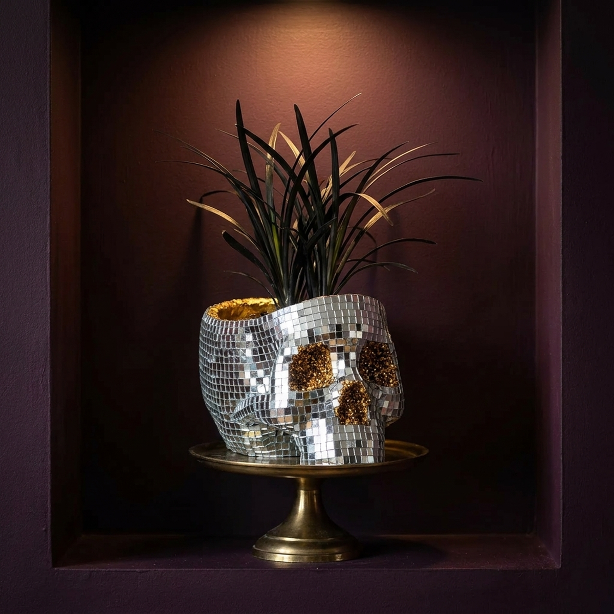 Disco skull planter with dark spiky foliage on a brass pedestal in a shadowed alcove