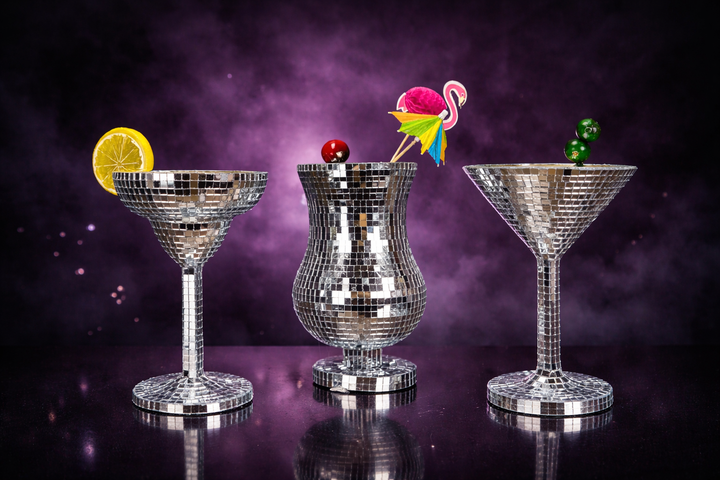 Three silver mirror mosaic cocktail glasses with colourful garnish details on a purple backdrop
