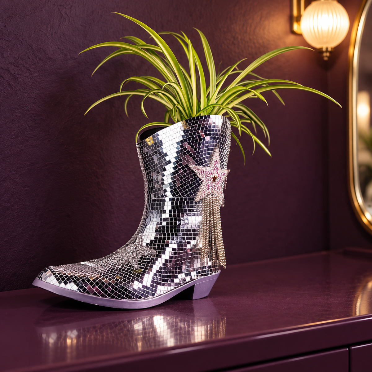 Silver mirror-tile cowboy boot planter with a star, chain fringe and spider plant on a plum dressing table.